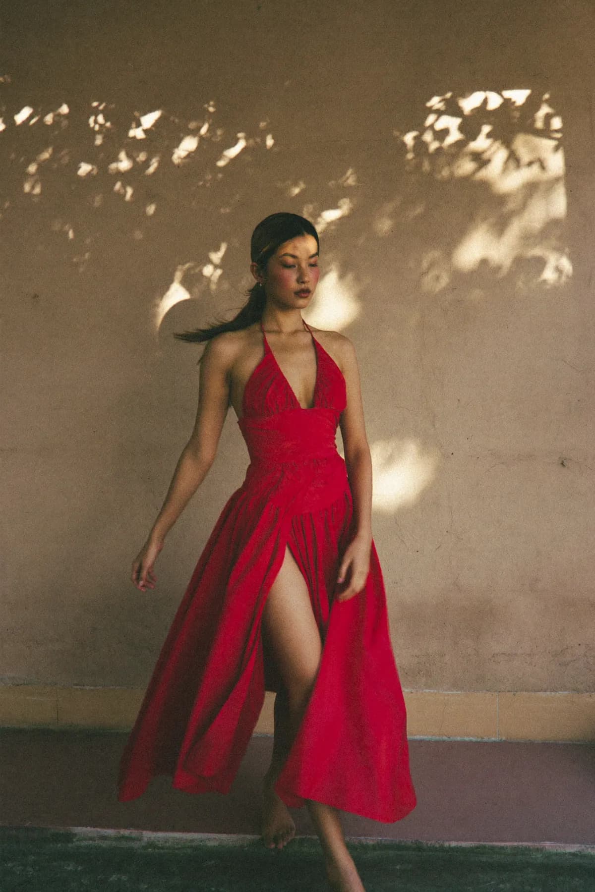 Woman in a red halter midi dress walking past a wall dappled with leaf shadows